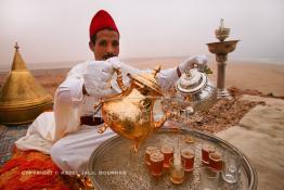 Image du Maroc Professionnelle de  Un marocain traditionnellement vêtu serve du thé à la menthe aux invités du grand Moussem de Tan Tan. Installée avec ses plateaux sur les hauteurs de la plage Chbika au environ de Tan Tan. Le Moussem est une sorte de foire annuelle, à la fois économique, culturelle et sociale. Il est classé par l'Unesco en 2005 comme patrimoine immatériel humain mondial. Chaque année la majorité des tribus et des grandes familles nomades du désert se retrouvent sur un site désertique pour célébrer ce grand Moussem où  danse, chants, course de chameaux et fantasia font partie des festivités. , Samedi 18 septembre 2004, ((Photo / Abdeljalil Bounhar) 